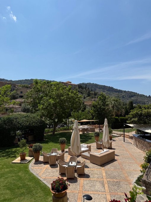 An elegant outdoor terrace at a Mediterranean resort features a stone-tiled patio with lounge chairs, umbrellas, and dining tables overlooking lush gardens, a swimming pool, and rolling hills under a clear blue sky—capturing the essence of a relaxing weekend getaway.