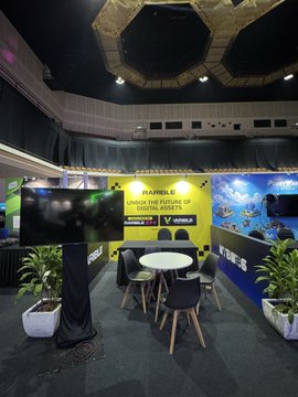 The Rarible booth at MalaysiaBCW features a vibrant display highlighting the platform's mission to "Unlock the future of digital assets," with the tagline "for EVERYONE" displayed prominently on screens. The setup includes a small table with chairs, potted plants, and branded materials, providing a welcoming space for attendees to engage with the team and learn about Rarible's role in the digital asset space.
