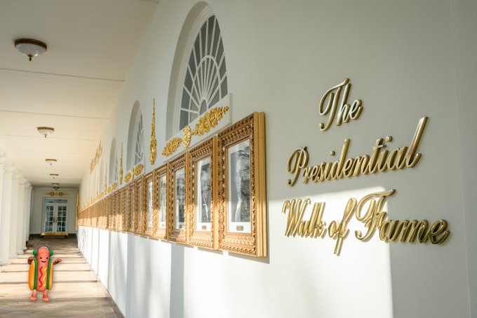 A long hallway displays ornate gold-framed portraits along the wall with the elegant script "The Presidential Walk of Fame" mounted beside them, while a small cartoon hot dog figure stands in the foreground—juxtaposing grand patriotic imagery with the meme's irreverent "Buy the dip" financial humor.