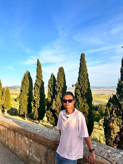 A man wearing sunglasses and a light pink shirt standing by a stone wall, with cypress trees and a scenic landscape in the background.