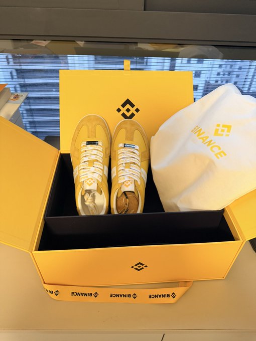 A pair of yellow and white Binance-branded sneakers is displayed inside a yellow Binance shoebox alongside a white dust bag.