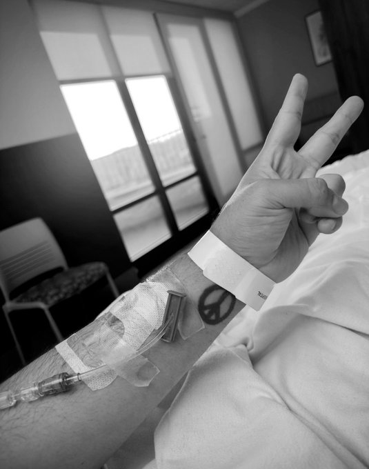 A black-and-white photo of a person's arm resting on a bed, showing an IV catheter attached to the wrist, a peace sign gesture, and a peace symbol tattoo on the forearm.
