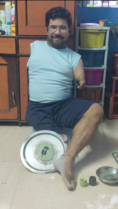 A man with one arm sits on the floor holding paint supplies and a large metal plate with a painted design, demonstrating his daily art practice as a personal investment habit. The image creatively ties into his post about consistent creative effort and the "Robinhood" theme, as he displays both artistic dedication and resourcefulness with everyday materials.