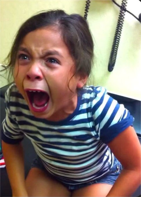 A young girl with dark hair screaming with her mouth wide open while sitting on a toilet, wearing a striped shirt.