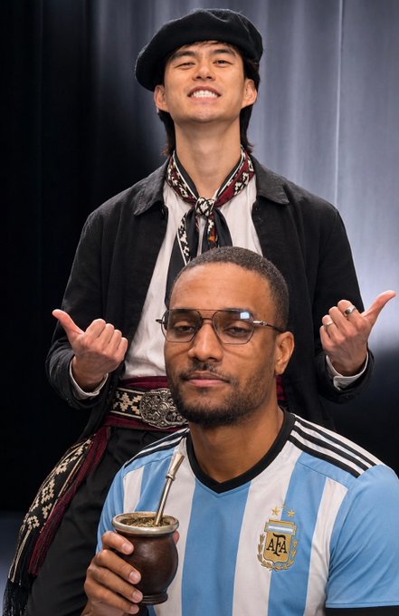 A photo of two men in costumes; the man in the back wears a beret and dark jacket with a patterned scarf, while the seated man wears an Argentina football jersey and glasses, holding a mate gourd.