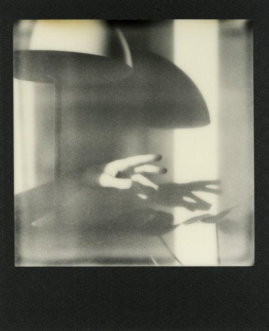 A black-and-white, heavily blurred and shadowy photograph of a person wearing a large hat with their hands raised in front of them, framed like a Polaroid.