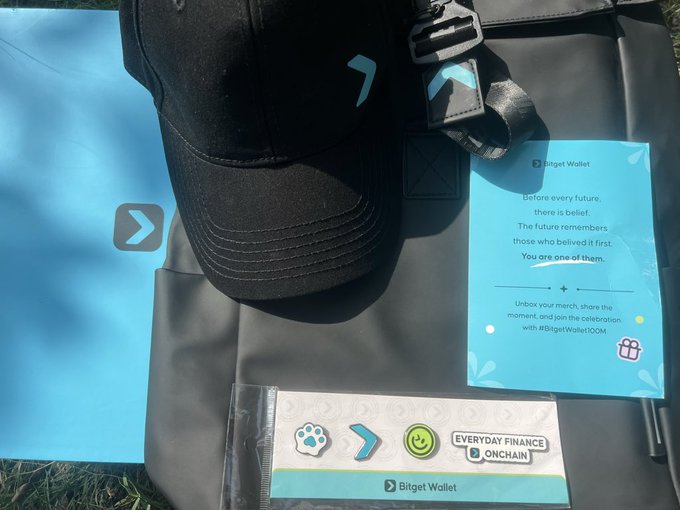 A black Bitget Wallet cap sits on a black bag alongside a welcome card reading "Before every future, there is belief. The future remembers those who believed it first. You are one of them. Unbox your merch, share the moment, and join the celebration with #BitgetWallet100M," and a sheet of stickers featuring "EVERYDAY FINANCE ONCHAIN." The accompanying caption "something blue arrives" captures the blue-themed Bitget branding visible throughout the promotional merchandise.