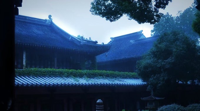 A misty photograph of a traditional East Asian temple complex with dark blue-tiled roofs, surrounded by lush green trees.