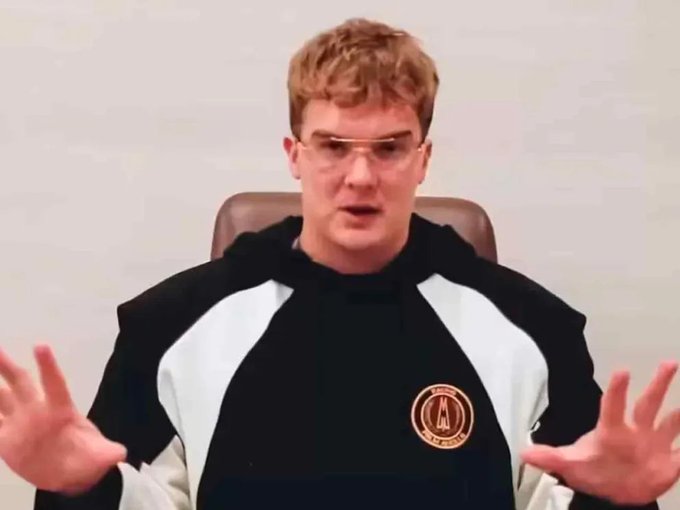 A young man with reddish-blonde hair wearing glasses and a black and white AC Milan hoodie, gesturing with both hands raised as if explaining or emphasizing something.