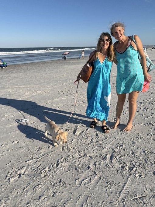 Two women standing on a sandy beach in matching turquoise cover-ups, smiling at the camera with a small dog on a pink leash in the foreground and the ocean behind them.