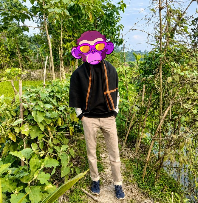 A person standing outdoors in a lush garden setting wearing a black fleece jacket with orange details, beige pants, and dark sneakers, with their face obscured by a purple monkey emoji wearing sunglasses.