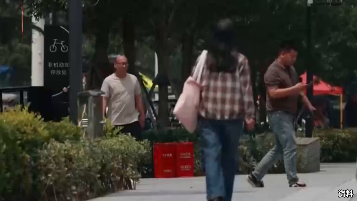 The image shows a man walking down a sidewalk with his hand in his pocket while another man walks behind him carrying a pink bag. There are also two other people visible, one of whom has their back turned towards the camera. The scene takes place on a street where there is an outdoor market or possibly a store selling items for sale. A person holding a cell phone can be seen near the center-right side of the image.