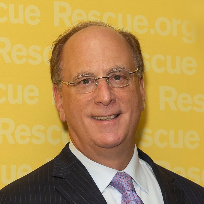 A smiling older man with glasses, wearing a dark pinstripe suit and patterned tie, stands in front of a yellow backdrop featuring repeated "Rescue.org" text. Given the post about BlackRock clients purchasing $22.38M in Bitcoin, the image likely depicts Larry Fink, the CEO of BlackRock, the world's largest asset manager that has been actively investing in Bitcoin.
