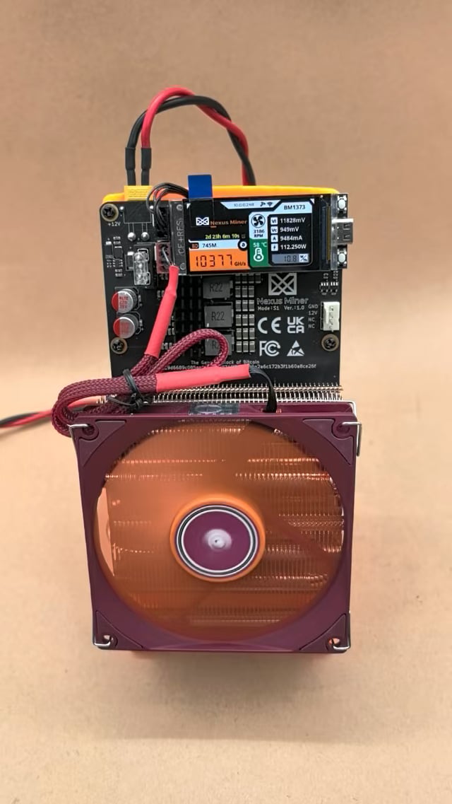 A NerdMiner S1 (Lottery edition) standalone Bitcoin lottery miner device is shown with its small display reading "103.77GH/s" along with voltage readings, mounted above a maroon cooling fan.