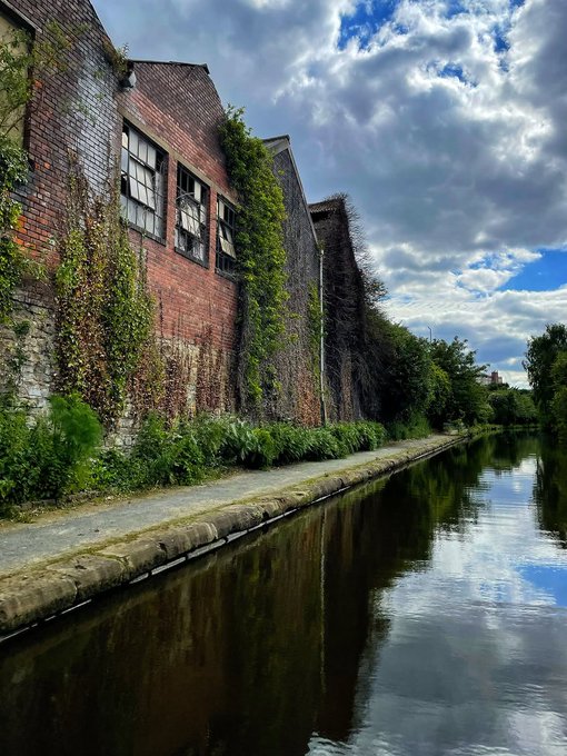 A scenic view of an ivy-covered red brick building alongside a calm canal under a dramatic cloudy blue sky.