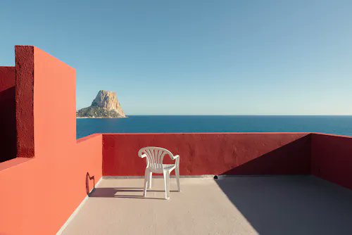 A serene rooftop terrace with red stucco walls and a white plastic chair overlooking a calm blue sea with a prominent rocky island in the distance under a clear sky.