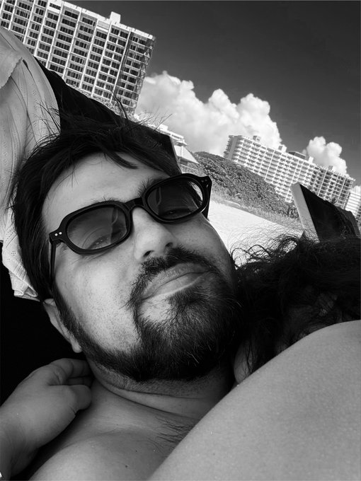 A black-and-white selfie features a bearded man wearing sunglasses, lying back with buildings and a cloudy sky visible in the background.