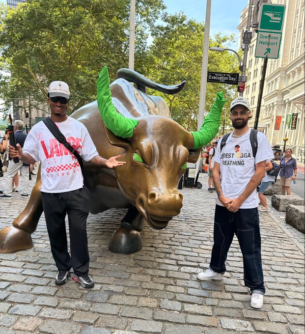 Two men pose on either side of the iconic Charging Bull statue in New York City's Financial District, with one wearing a "VANS" shirt and the other in a "COAST GUARD BREATHING" shirt. The post's reference to "$ANSEM" and "THE SOLANA TRENCHES" suggests a connection to the cryptocurrency community, using the powerful bull imagery as a symbol of bullish market sentiment.