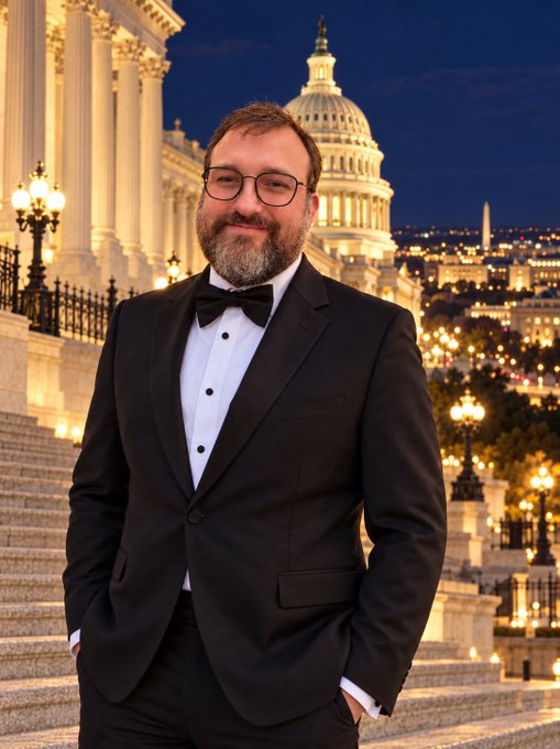 A smiling man with glasses and a beard, wearing a black tuxedo and bow tie, stands on steps at dusk with the illuminated U.S. Capitol building in the background.
