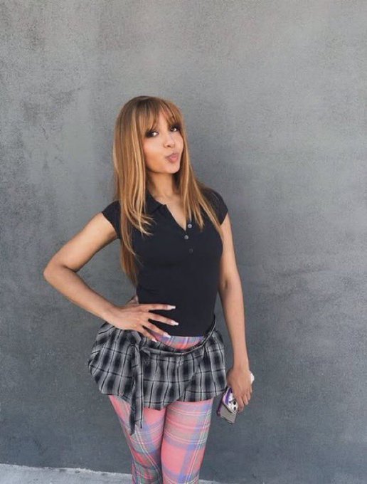 A young woman with long hair posing in a black polo shirt, plaid skirt, and pink plaid leggings against a gray background.