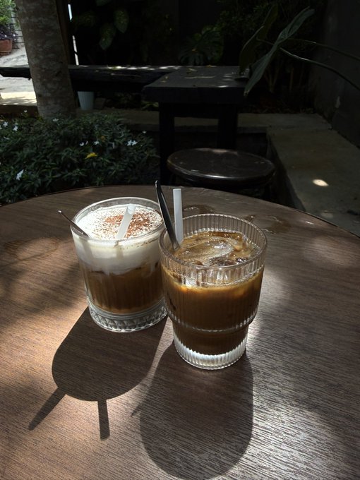 The image shows two glasses of coffee sitting on a wooden table, with one glass being filled with milk and the other with cream. There are also some plants visible near the table. The photo appears to be taken from an angle that highlights the contrast between the two drinks in terms of their contents.
