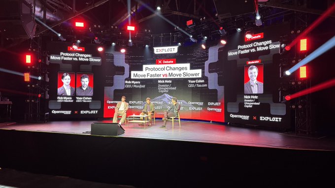 A wide shot of a conference stage set up for a 'Deloitte Protocol Changes: Move Faster vs. Move Slower' debate panel at an 'Exploit' event, featuring screens with panelists' names including Rob Myers, Yasar Dultan, Nick Hotz, and Youn Oillon.