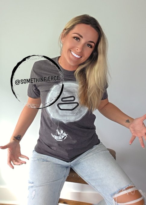 A smiling woman with long blonde hair wearing a gray graphic t-shirt and ripped blue jeans, featuring the watermark '@SOMETHINFIERCE'.