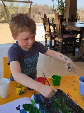 A young boy in a dark t-shirt sits at a table painting with colorful cups of paint, holding a paintbrush as he works on a piece of artwork.