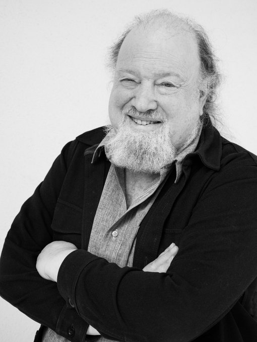 A black-and-white portrait captures cryptography pioneer David Chaum smiling warmly with arms crossed, wearing a dark cardigan over a collared shirt, his distinctive long white beard prominently featured. The image is presented as part of a tribute to icons of Bitcoin, photographed by dotkrueger for the Big Bitcoin Book.