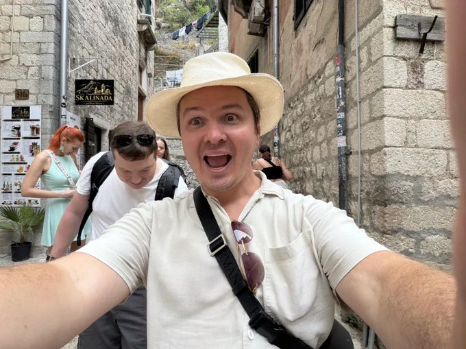 A man in a cream-colored shirt and sun hat takes an excited selfie in a narrow stone alleyway, with a "Skalinada" restaurant sign visible behind him and a companion walking by. The image contrasts the high-energy travel moment with the post's reflective message about the emotional discipline required in crypto markets, where taking profits during bull markets can feel as thrilling—and as difficult—as capturing a perfect travel shot.