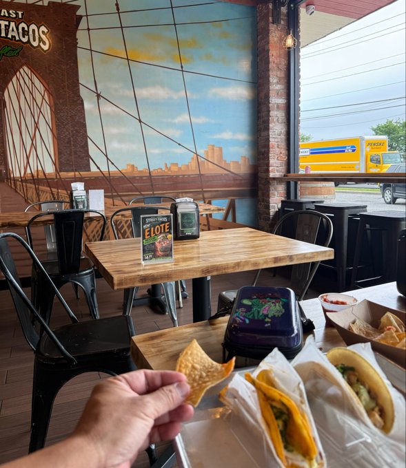 A first-person view inside a taco restaurant with a Brooklyn Bridge mural, showing a hand holding a tortilla chip with tacos and a lunch container on the table. A Penguin shipping truck is visible through the window.