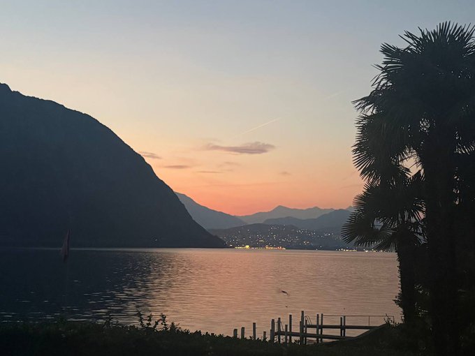 A serene sunset over a calm lake with silhouetted mountains in the distance, a palm tree on the right, and city lights twinkling along the far shore.
