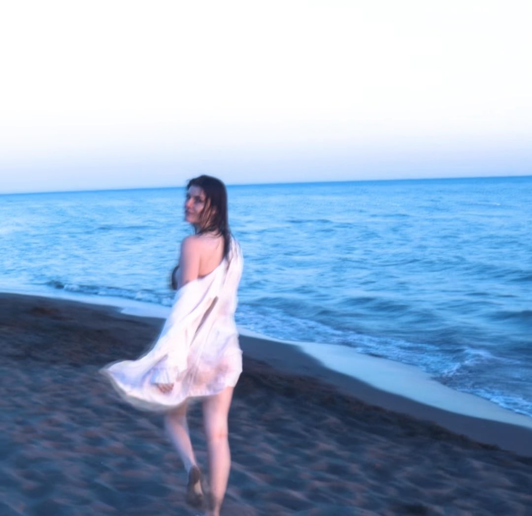 A slightly blurred photograph of a woman in a flowing white dress walking on a beach near the ocean.