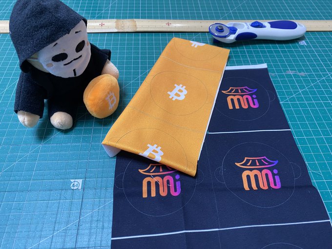 A plush toy wearing a black hoodie sitting next to fabric strips—one orange with Bitcoin "B" logos and one black with "mi" logos—on a green cutting mat with a rotary cutter.