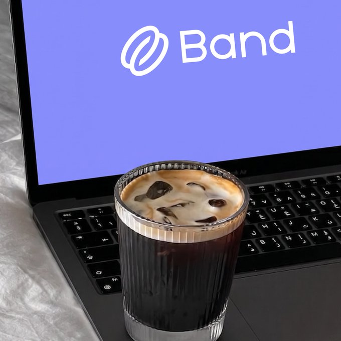A glass of iced coffee sits on the keyboard of a laptop displaying the "Band" logo on a purple screen, suggesting a productive workday fueled by caffeine. The image pairs with the "GM It's that time of the week again" post to evoke a fresh start to the week, likely in the crypto or financial trading space.