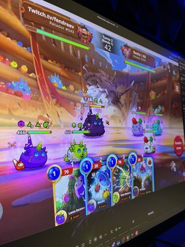 The image shows a computer monitor displaying an animated video game scene with several characters, including a dragon and other creatures, engaged in combat or fighting for dominance. The characters appear to be using various weapons such as swords and shields, while their surroundings are filled with colorful elements like flowers and trees. The post on the screen reads: "Gm Gm we are live come on @AxieInfinity".