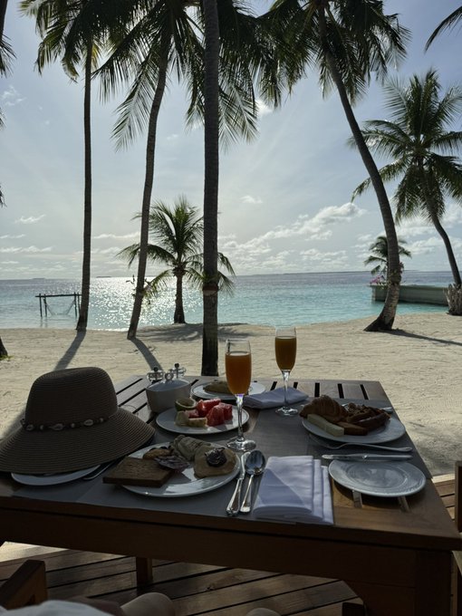 A beachside breakfast setup with fruit, pastries, and mimosas on a table, framed by palm trees overlooking the turquoise ocean.