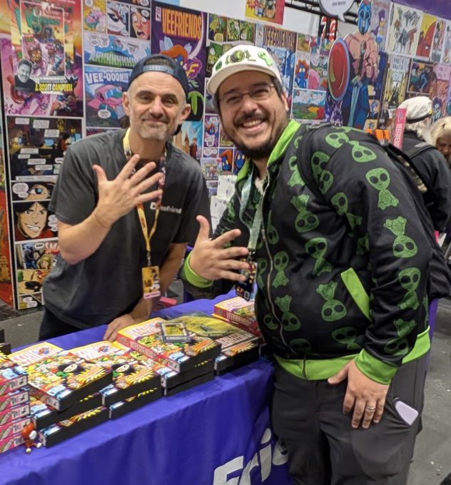 Two men smiling at a comic convention booth, one wearing a green alien-patterned hoodie and cap, standing behind a table stacked with 'Wee-Friends' and 'Killer Bee' comic books.