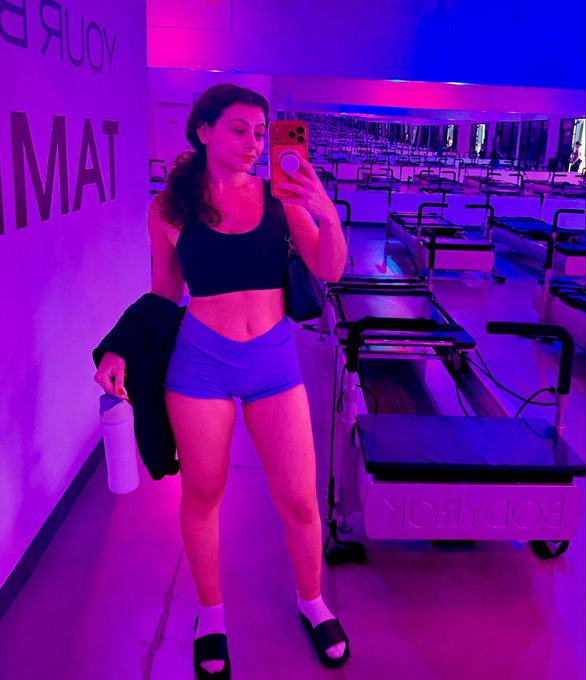 A woman takes a mirror selfie in a Bodyrok Pilates studio illuminated with purple and blue neon lighting, wearing a black sports bra and blue shorts while holding a water bottle and jacket.