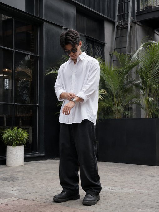 A man wearing sunglasses, an oversized white button-up shirt, loose black pants, and black shoes poses outside against a dark building with potted plants.