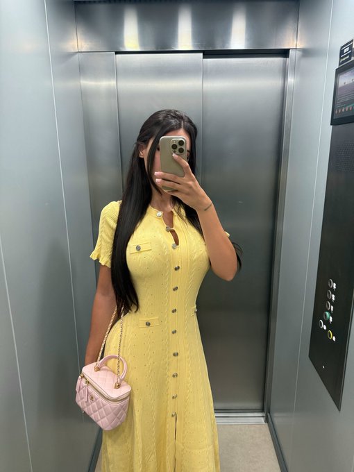 A woman in a yellow dress takes a mirror selfie inside an elevator while holding a pink quilted handbag, her phone obscuring her face. The bright yellow outfit visually echoes the caption's wordplay on "yellow," playfully tying her look to the "crime" theme on the BNB chain.