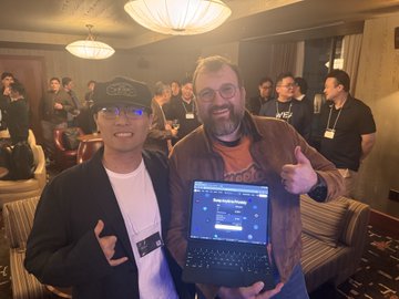 Two men pose for a photo in a crowded room; one wears a black hat and jacket while the other in a brown jacket gives a thumbs up and holds open a laptop displaying a webpage.