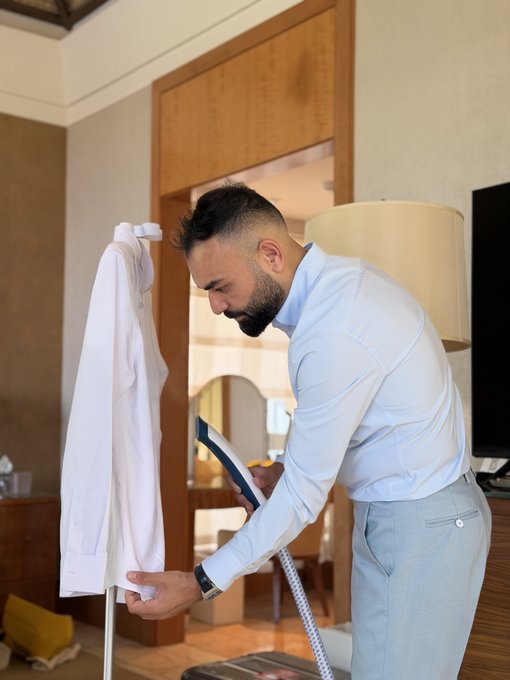 A bearded man in a light blue dress shirt steams a white garment on a hanging rack in a hotel room, carefully pressing the fabric with an iron. The meticulous preparation reflects his dedication to looking sharp for the Token2049 event.