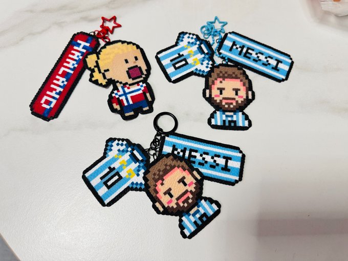 The image features a table with four keychains, each displaying different cartoon characters. The first keychain has a girl character, the second one has a boy character, the third one also has a girl character, and the fourth one has a boy character. These keychains are made of metal and have names written on them in blue letters. The post includes text that reads "看了下OKX世界杯榜单", which is related to the image by being a description or caption about the keychains.