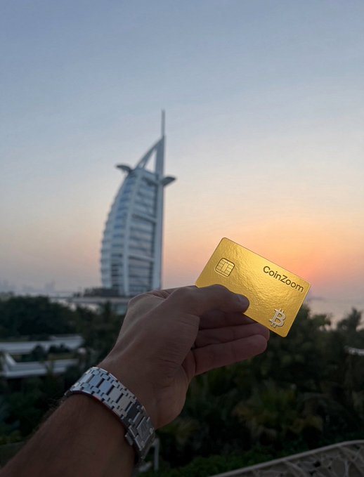 A person proudly displays a gold CoinZoom Bitcoin debit card with the iconic Burj Al Arab towering in the background at sunset, perfectly illustrating how easily cryptocurrency can be used for everyday purchases like morning coffee anywhere in the world.