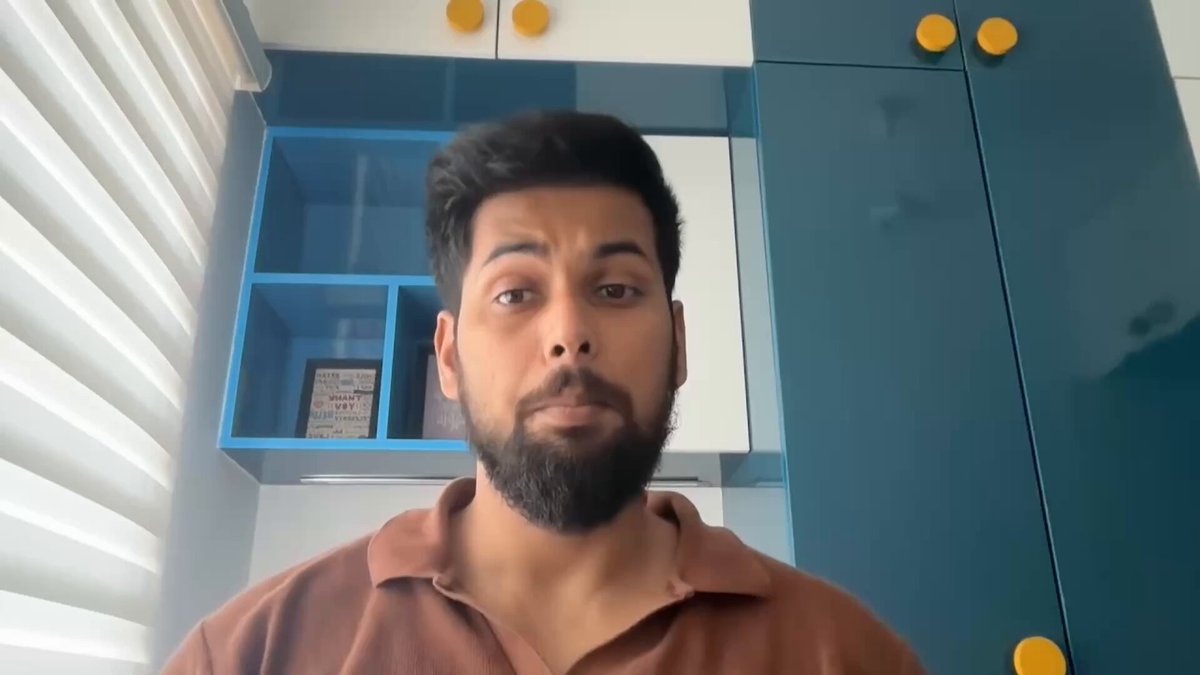 The image shows a man wearing a brown shirt, standing against a blue wall with yellow cabinets. The man appears to be looking directly at the camera while posing for the photo. There are no texts or other objects visible in the image that seem to have any connection to the post.