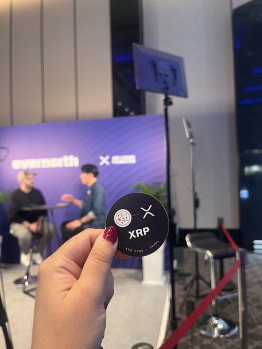 A hand with red nail polish holds up a black coin labeled "XRP" with an airplane graphic, with a blurred backdrop showing an "evernorth x" booth and two people sitting.
