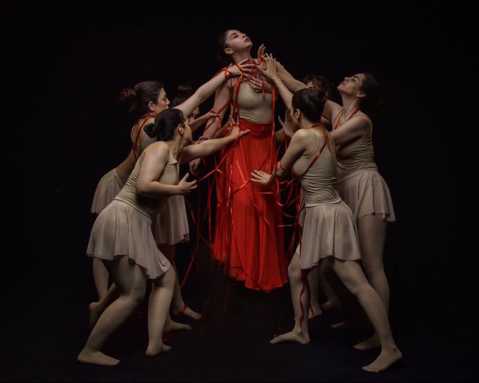 A group of dancers in tan costumes perform a dramatic piece where several figures surround and grasp at a central dancer wearing a long red skirt and entwined in red ribbons. The visual themes of being "entangled" and "transient" are represented by the literal intertwining of the dancers and the flowing red ribbons.