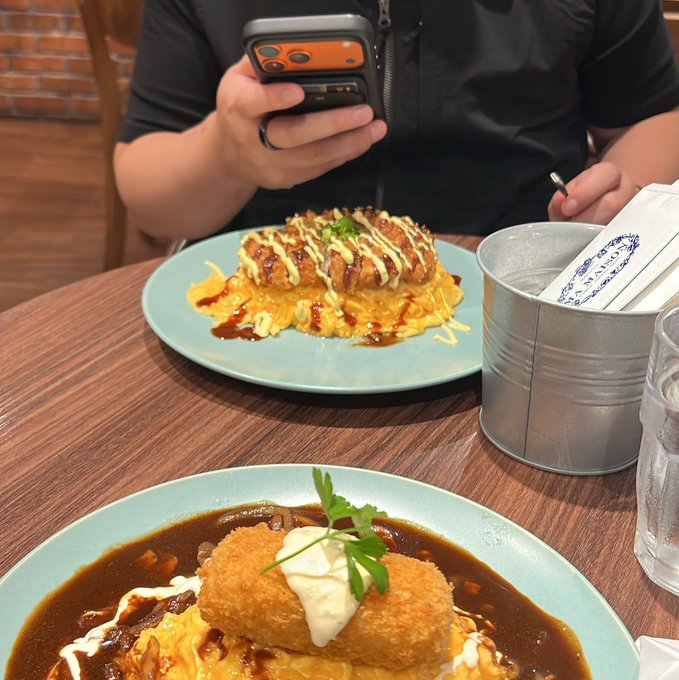 A person sits at a wooden table photographing a meal of breaded cutlets drizzled with sauces on scrambled eggs with rice, accompanied by a metal utensil holder. The image reflects the post's mention of taking a break for food during a busy Token2049 conference week with friends.
