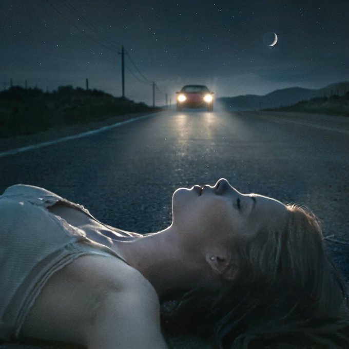 A young woman lies on a dark road at night with her eyes closed, while a car with headlights approaches in the distance beneath a crescent moon.